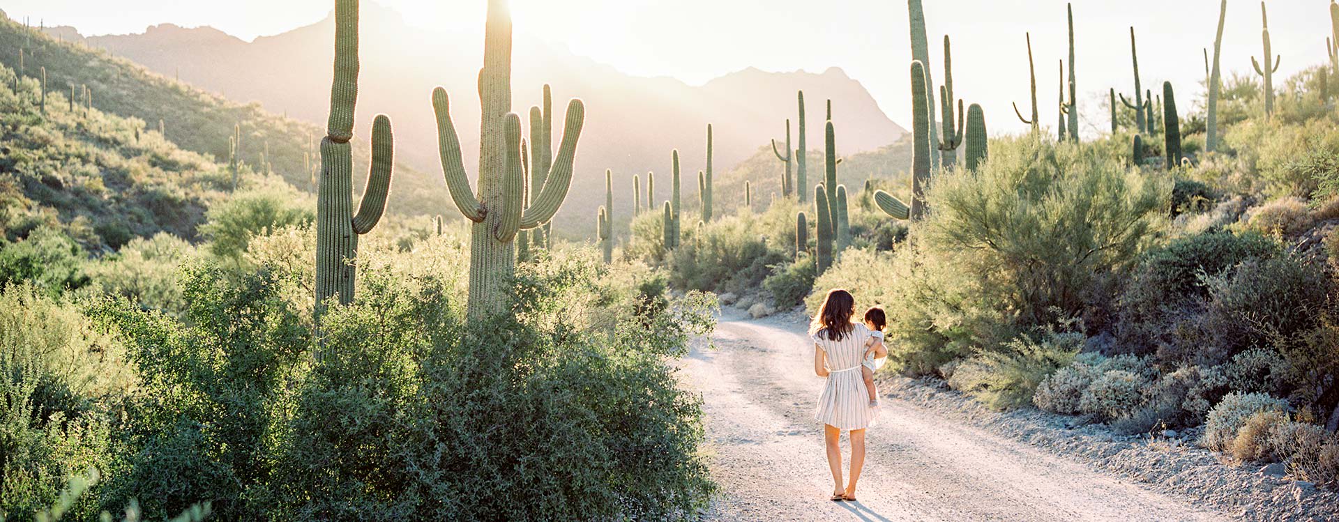 A woman holding her baby walk down a desert trail toward the sunlight